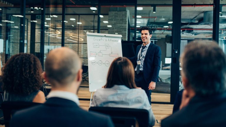 A confident leader standing in an office environment, surrounded by engaged team members who are naturally gravitating toward them. The lighting should emphasize the leader's calm, assured posture.