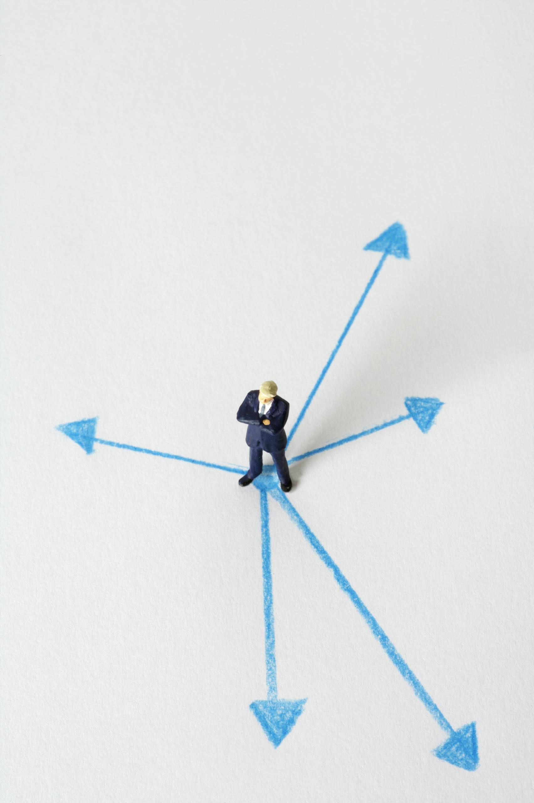 Miniature businessman stands at crossroads of blue arrows on white background, symbolizing choices.